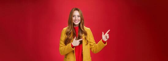 Cheerful friendly-looking and energized helpful redhead girl in yellow coat showing way pointing at upper left corner and smiling broadly with satisfied grin posing delighted over red background photo