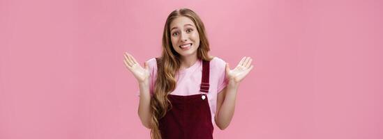 Sorry I have no idea. Silly unaware cute friendly-looking young female student in dungarees over t-shirt shrugging with palms raised in surrender smiling awkward as if being clueless over pink wall photo