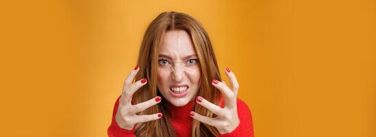 Close-up sot of pissed and angry redhead woman raising hands and clenching them with anger and outrage being irriated and annoyed grimacing from dislike, hate and pressure over orange wall photo