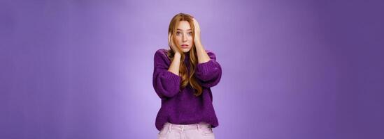 Girl in stupor feeling shocked and troubled as realising huge mistake made, holding hands on face and head popping eyes at camera confused and hopeless, posing anxious over purple background photo