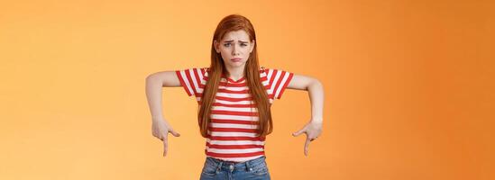 Cute timid hesitant ginger female taking tough decision feel pressured upset, frowning, pulling gloomy unhappy face, pointing down disappointed, uneasy taking decision, orange background photo