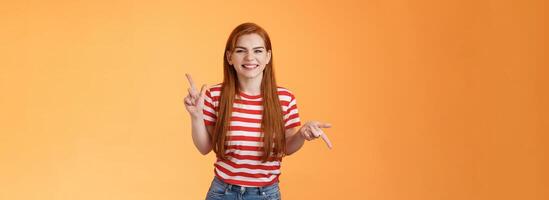 Carefree daring redhead cheerful girl dancing, pointing sideways, showing different sides, left right copy space, laughing and smiling broadly, joyfully recommend cool product, orange background photo