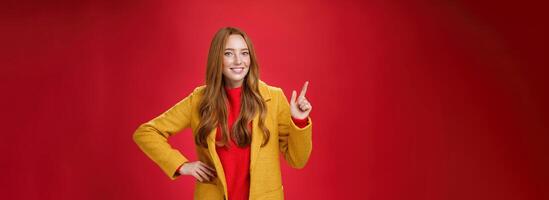 Charming young female in yellow coat holding hand on waist and pointing at upper left corner, smiling broadly as showing or promoting interesting proposal over red background, in good mood photo