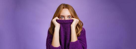 Redhead girl scared winter is comming hiding in collar of warm sweater popping eyes shocked and stunned at camera as if frightened and insecure standing terrified over violet background photo
