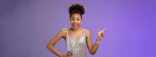 Smiling elegant african-american girl prom party wearing silver glittering evening dress pointing left index finger widen eyes surprised impressed receive opportunity lifetime, blue background photo