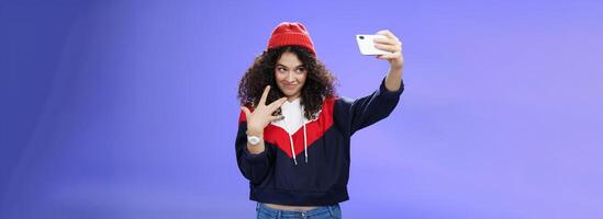 Cool and stylish good-looking caucasian female with curly hair in trendy red beanie and sweatshirt looking from under forehead awesome and swah taking selfie with smartphone holded in extended arm photo