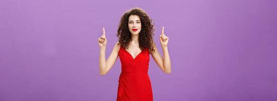 Feeling on top after making stylish makeup. Cute and tender glamourous european female in red evening dress and lipstick with dark curly hair smiling gently and pointing up with raised hands photo
