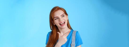 Sassy good-looking dreamy redhead female student contemplate cool performance, tilt head joyfully pointing look left, discuss holiday plans, curiously asking question, posing blue background photo
