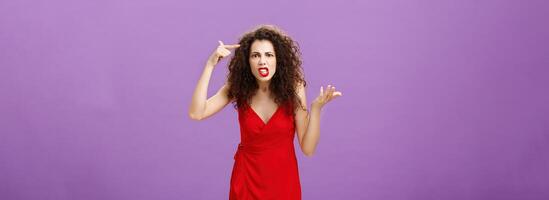 Outraged and pissed wife critisize dumb idea, outraged and fed up with stupid husband actions rolling finger near temple gesturing with raised palm, being angry and questioned over purple background photo