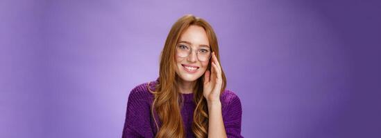 Smart and creative good-looking female book lover with ginger hair in glasses smiling with friendly gaze at camera, touching face shy and tender posing delighted and carefree against purple background photo