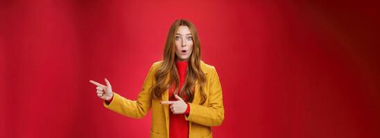 Speechless questioned and confused surprised redhead girl in yellow autumn coat pointing right open mouth curiously and staring at camera puzzled and clueless waiting explanation over red wall photo
