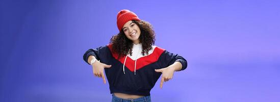 Waist-up shot of cool and stylish young woman with curly hair smiling flirty and joyful pointing down showing promotion as tilting head happily and posing over blue background in outdoor clothes photo