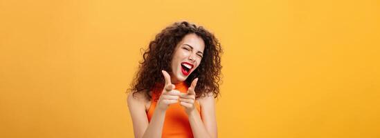 Hey what is up. Friendly and outgoing attractive curly-haired female with red lipstick pointing at camera with finger guns as if greeting or hinting friend winking at camera over orange background photo