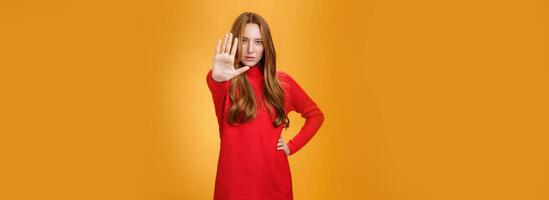 Confident sensual and attractive redhead female with self-assured gaze extending palm towards camera in stop and prohibition gesture demanding halt, stand still over orange background photo