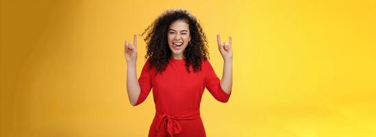 Cute excied and daring woman in 25s sticking out tongue as fooling around having fun on concert winking joyfully and showing rock-n-roll gesture feeling amused and alive over yellow background photo