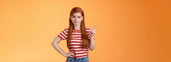 Doubtful redhead girl not buy your talks, smirking hesitant express disbelief, unimpressed about suspicious offer, pointing upper left corner wondered, stand orange background uncertain photo