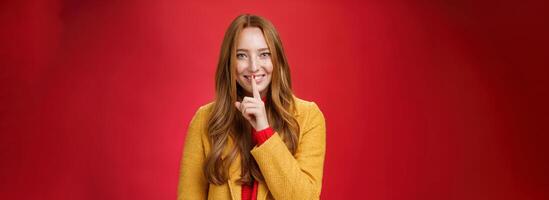 Shh keep it secret. Flirty and gentle attractive ginger girl with freckles teeling mysterious things showing shush gesture with index finger on mouth, smiling having surprise over red background photo