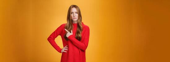 Girlfriend asking serious question being bothered by unfamiliar woman on family dinner pointing behind frowning and squinting suspicious expressing disbelief, doubt and vigilance over orange wall photo