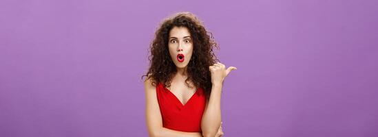 Curious and questioned amazed woman with curly hairstyle in stylish red dress folding lips in wow sound pointing left with thumb asking question about stunning and thrilling scene over purple wall photo