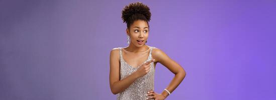 Curious unsure african-american attractive elegant young woman in silver glittering evening dress look pointing left intrigued discuss strange person gaze doubtful uncertain, blue background photo