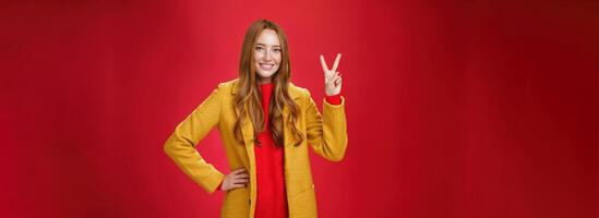 Portrait of happy playful and cute redhead woman with freckles in yellow coat holding hand on waist confident and self-assured showing peace or two sign delighted, beating weather conditions photo