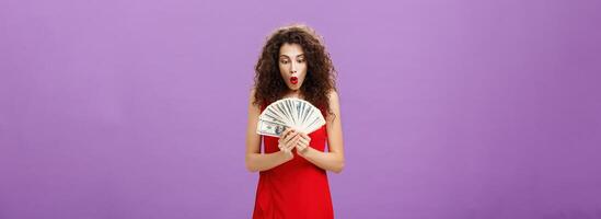 Portrait of surprised and amazed speechless woman folding lips making wow sound while holding and looking at lots of money being rich and successful receiving lots of cash over purple background photo