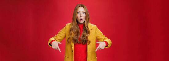 Impressed girl cannot believe what sees open mouth from surprise and excitement popping eyes questioned at camera, pointing down with index fingers asking question and being astonished over red wall photo