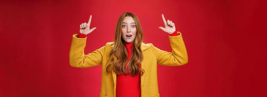 Surprised redhead girl in yellow coat open mouth impressed and thrilled as raising hands and pointing up telling about cool promotion with excited expression against red background photo