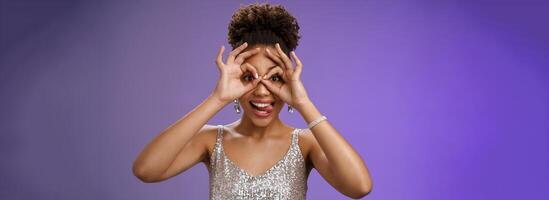 Playful funny good-looking african woman in afro hairstyle in silver glittering dress having fun show tongue smiling broadly make okay ok gesture on eyes look excited, standing blue background photo