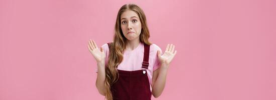 I am not involved. Portrait of unaware and clueless innocent cute woman raising palms in surrender pulling lips down as if being confused and uncertain what happening having no idea over pink wall photo