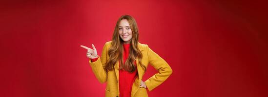 Stylish and self-assured attractive redhead woman with freckles in yellow outdoor coat pointing right with finger pistol and smiling joyfully as camera as showing cool promotion over red wall photo