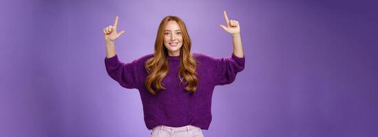 Scroll up and see cool copy space. Portrait of happy attractive and friendly young ginger woman with freckles in warm purple sweater raising hands pointing upwards as showing place photo