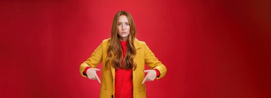 Portrait of sad and displeased moody girlfriend asking opinion with concerned upset expression pointing down looking focused and serious with unhappy face at camera over red background photo