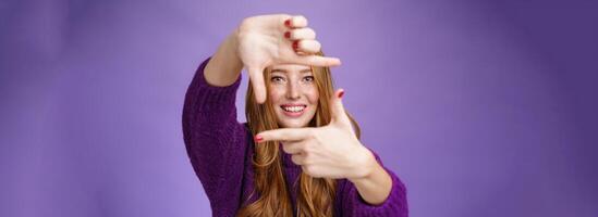 Imagine if. Portrait of hopeful and carefree creative charming redhead 20s woman showing frame with pulled hands and smiling through it as picturing bright and happy future over purple background photo
