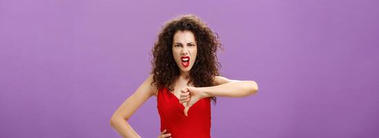 Bullshit I hate your lame ideas. Displeased and pissed lady boss with curly hairstyle in red evening dress growning grimacing from hate and anger showing thumbs down in rejection and disapproval photo