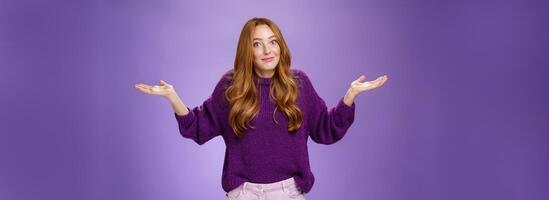 Well who cares, nothing to do. Portrait of funny and careless unbothered chill redhead female shrugging with spread hands and tight smile being unaware and not giving interest over purple background photo