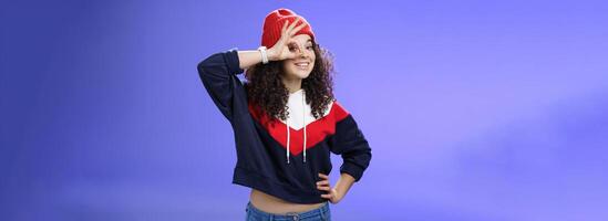 Okay girl agrees. Friendly-looking happy and charming young curly-haired girl in warm beanie showing ok gesture or circle over eye as peeking at camera and smiling broadly with joy over blue wall photo