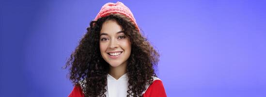 Headshot of tender and cute curly-haired 20s woman in warm beanie and cool sweatshirt smiling broadly enjoying awesome sunny and chilly days outdoors having fun posing over blue background photo