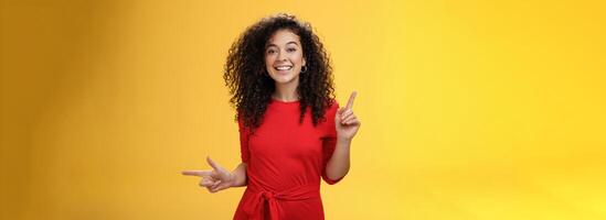 Charismatic cute and happy young curly-haired european female in red dress dancing and pointing up and right, showing sideways as presenting choices picking, making decision and smiling at camera photo