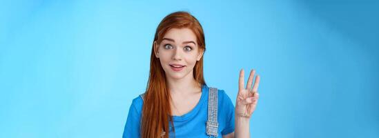 Surprised cute redhead caucasian girl show number three, third, counting fingers, raise eyebrows ambushed, smiling curiously, stand blue background playful tender expression photo