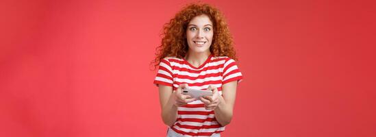 Excited happy cute redhead curly-haired girlfriend eager start learn how play boyfriend game look amused cheerful camera smiling joyful hold horizontal smartphone load app red background photo
