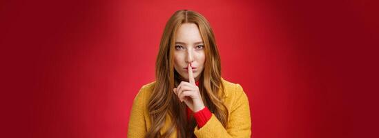 Shh keep out secret safe. Portrait of sensual attractive and daring redhead female in yellow coat showing shush gesture with index finger on lips, squinting looking determined and sassy over red wall photo