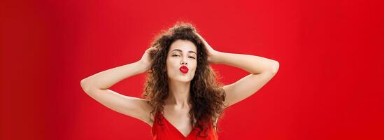 Waist-up shot of confident flirty and elegant young rich woman with curly hairstyle in red lipstic and evening dress touching hair folding lips in mwah giving kiss to admirer feeling self-assured photo