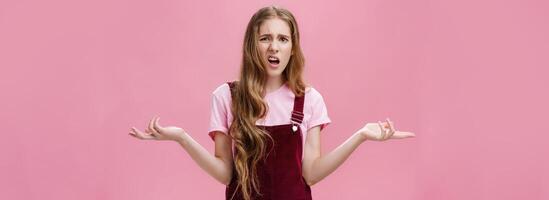 So what, no big deal. Rude and impolite arrogant young girl with high ego in overalls spreading hands sideways in questioned, bothered gesture expressing confusion, feeling pissed with stupid question photo