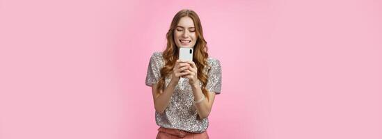 Charming glamour sociable stylish caucasian female using smartphone looking mobile phone screen look gadget display amused writing post upload photo social web page texting, pink background