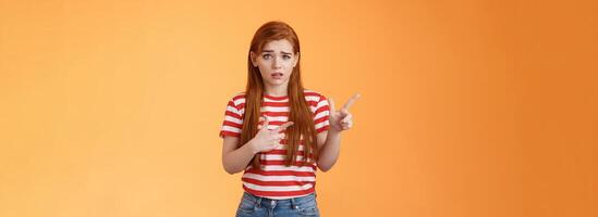 Insecure questioned frustrated cute redhead girl feel nervous unsure about suspicious offer frowning, grimacing doubtful and hesitant, pointing left look camera puzzled, asking question photo