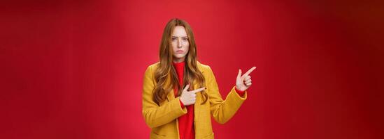 Displeased offended and gloomy girlfriend asking question, pouting and frowning from insult pointing at upper left corner disappointed and upset posing dissatisfied against red background photo