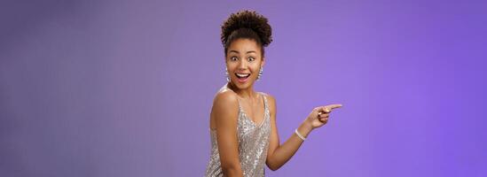 Impressed elegant good-looking african girl glittering shiny evening dress pointing index finger left turn camera wondered amused smile delighted impressed magian trick during show, blue background photo