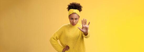 Brave serious-looking female african american femenist look from under forehead confident powerful pose raise palm stop enough not decline gesture forbid behave bad, standing yellow background photo
