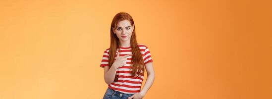 Click right now. Confident sassy good-looking redhead woman smiling cunning self-assured, pointing left, showing copy space promo, grinning empowered, know what best, orange background photo